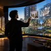 Stand de vendas imobiliário com maquete iluminada, telas interativas e cliente explorando plantas de apartamentos.